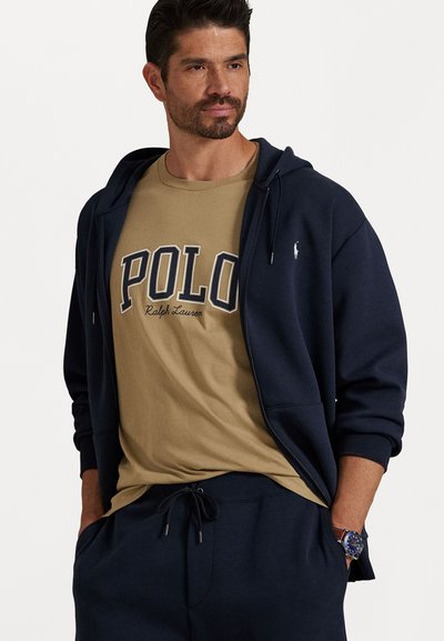 Moški nosi mornarsko zadrgo, tan majico polo Ralph Lauren, mornarske hlače s vrvico in modro uro na zapestju, roke v žepih.