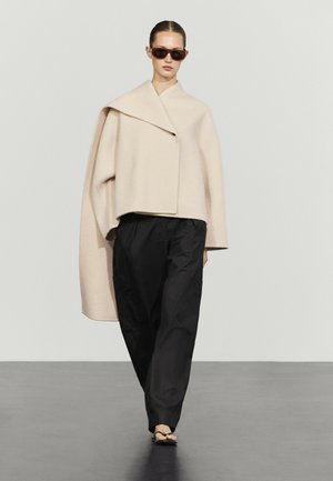 Frau mit zurückgekammtem Haar, die große braune Sonnenbrillen, eine beige übergroße asymmetrische Jacke, schwarze weite Hosen und schwarze Sandalen trägt.
