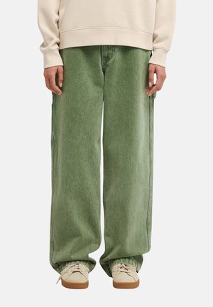 Persona che indossa jeans verdi larghi, felpa color crema e sneakers beige, in piedi contro uno sfondo chiaro e uniforme. È visibile solo la parte inferiore del corpo.