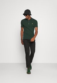 Mørk grønn polo skjorte med en gul logo, svarte bukser, grønne sko og en svart bøttehatt. Modellen går, iført solbriller.
