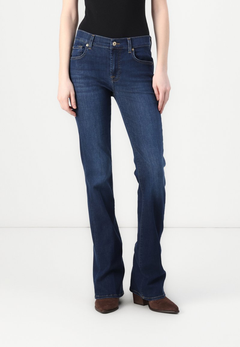 Jeans in denim blu scuro con design a campana, dotati di tasche frontali, chiusura con bottone e zip, e leggera scoloritura vicino alle ginocchia.