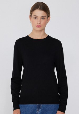 Junge Frau mit braunem, zurückgebundenem Haar, die kleine Creolen, einen schwarzen Pullover mit Rundhalsausschnitt und langen Ärmeln und blaue Jeans trägt, steht vor einem schlichten Hintergrund.