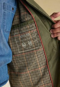 Rokas turēt atvērtu olīvu zaļas krāsas jaku ar brūnu un melnu pārbaudes oderi, kurā iekšējā kabatā ir "Joules" birka.