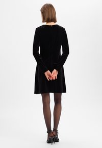 Schwarzes Samtkleid mit langen Ärmeln, rundem Ausschnitt und Reißverschluss auf der Rückseite. Über-kurzer Länge und ausgestelltem Rock, kombiniert mit durchsichtigen schwarzen Strumpfhosen.
