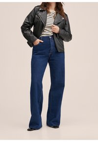 Veste en cuir noire portée sur un haut rayé, associée à un jean bleu foncé taille haute à jambes larges. Chaussures noires à bouts pointus.