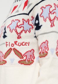 Pletený sveter s krémovým pozadím, farebnými vzormi vrátane červených sŕdc a modrých tvarov, s nápisom "Fiorucci" vyšitým červenou farbou.
