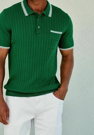 Homme portant un polo vert côtelé à manches courtes avec col et poche bordés de blanc, associé à un pantalon blanc, main dans la poche.