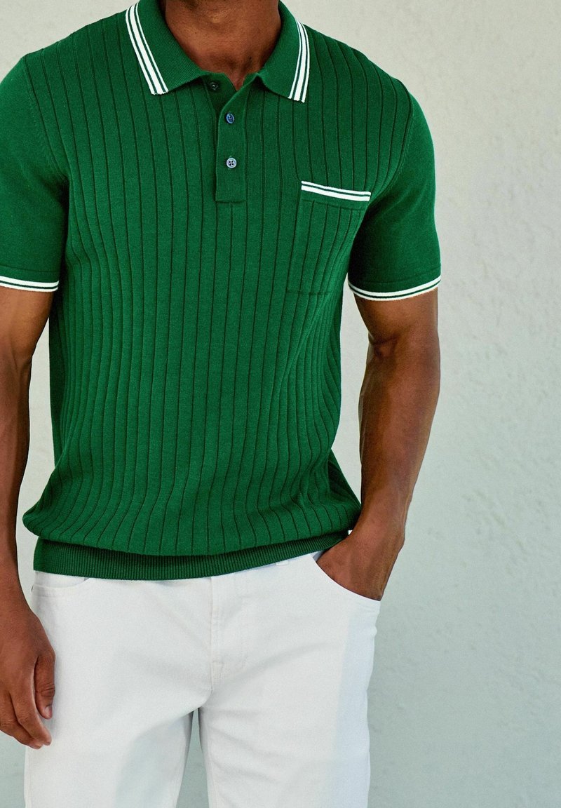 Homme portant un polo vert côtelé à manches courtes avec col et poche bordés de blanc, associé à un pantalon blanc, main dans la poche.