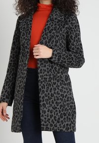Kabát s leopardím potiskem v šedé a černé barvě, vyrobený z měkkého materiálu, s klopovým límcem a dvěma předními kapsami, stylizovaný přes červený top.
