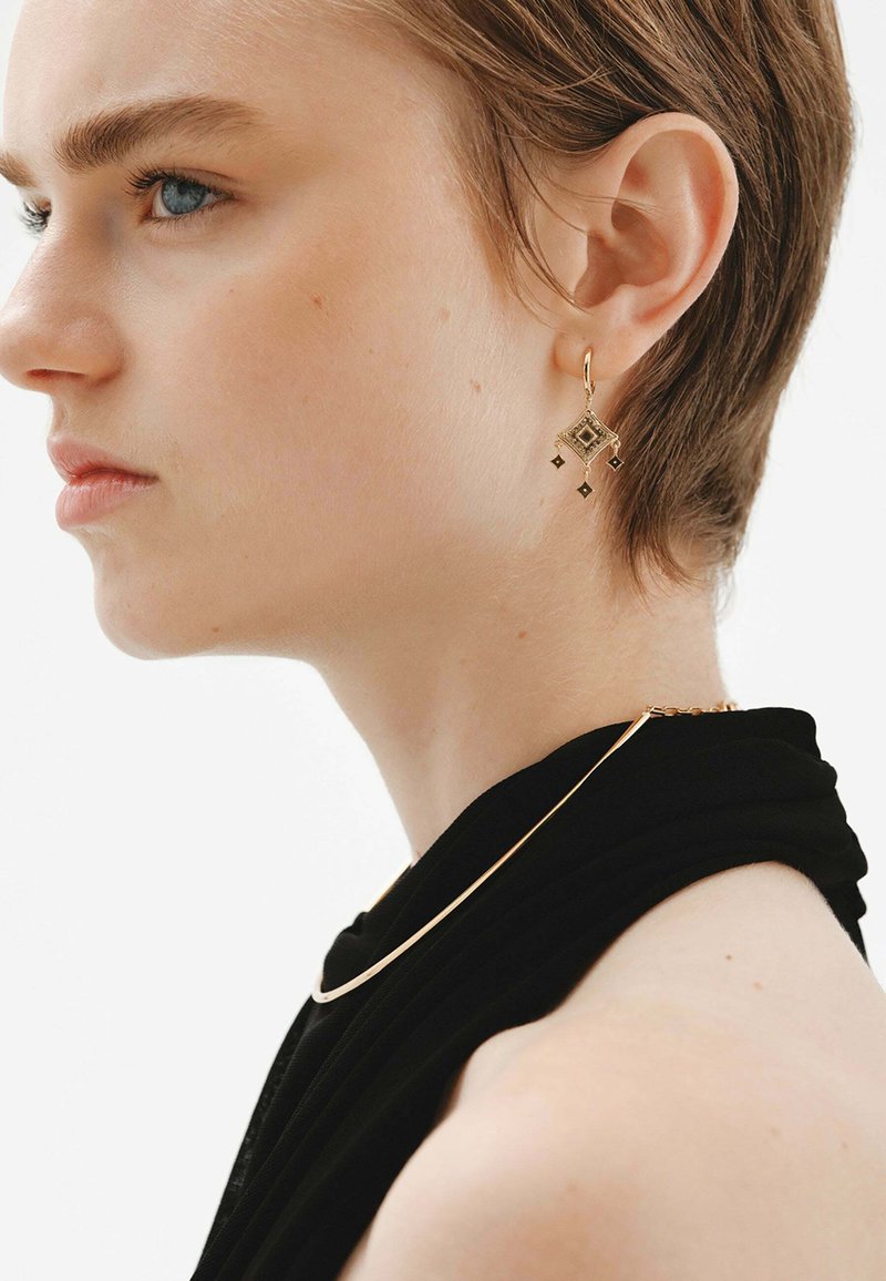 Pendientes geométricos en tono dorado con formas de diamante y acentos colgantes, combinados con un collar de cadena dorada minimalista y una blusa de tela negra.