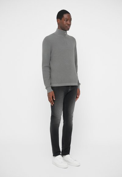 Grey, textured pullover with a high collar and half-zip closure, paired with dark jeans and white sneakers, standing against a plain background.