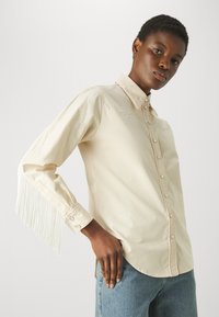 Crèmekleurig shirt met lange mouwen, franjes aan de linkermouw en geborduurde accenten op de borst. Knopen aan de voorkant.