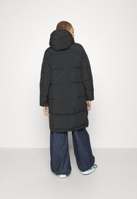 Zwarte gewatteerde jas met een capuchon, voorzien van horizontale stiksels, lange mouwen en een middellange snit. Gecombineerd met losse spijkerbroeken en sneakers.