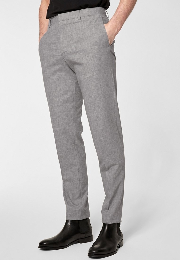 Homme portant un pantalon habillé gris clair slim fit avec poches latérales et bottines en cuir noir, debout les mains dans les poches.