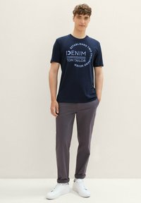 Námořnické bavlněné tričko s bílým kruhovým textem a logem. V kombinaci se šedými kalhotami a bílými teniskami, stojí před neutrálním pozadím.