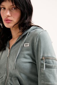 Jonge vrouw met donker golvend haar in een vervaagde groene hoodie met rits en een "JUICY" patch, met een gebreeide zak op de mouw.