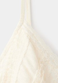 Gros plan d’une lanière de soutien-gorge en dentelle crème et d’une coque en tissu texturé avec des détails en dentelle florale sur un fond clair.