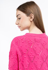 Maglione rosa lavorato a maglia con un motivo a diamante testurizzato, caratterizzato da accenti a pompon in rilievo e un collo a costine. I punti visibili creano un design dettagliato.