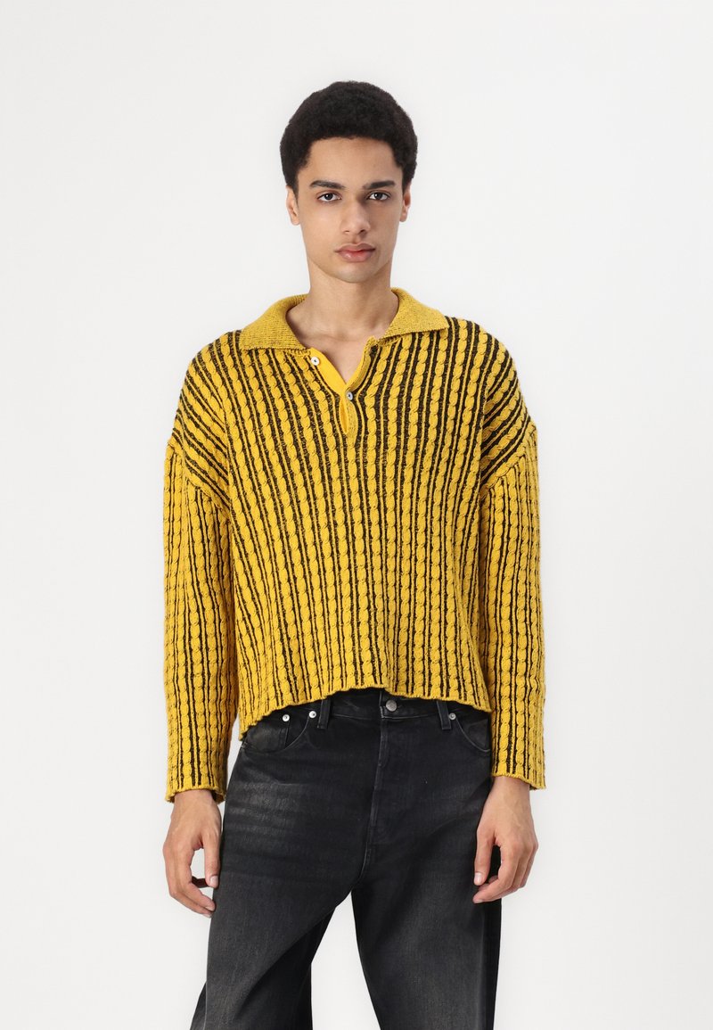Pull en tricot rayé jaune et noir avec col, fermeture à boutons sur le devant et coupe courte. Surface texturée avec des lignes côtelées.