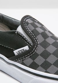 Scarpa slip-on a scacchi neri e grigi con cuciture bianche, etichetta con marchio e tomaia in tessuto texture; presenta una suola in gomma e dettagli laterali.