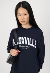 Tamsiai mėlyna sportinė palaidinė su baltais užrašais "KNOXVILLE Leisure Club EST. 1982", platus kirpimas, apvalus iškirpimas ir per didelės rankovės. Juoda petnešų kuprinė.