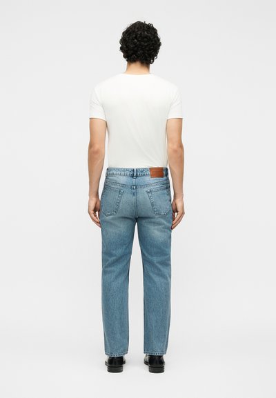 Άνδρας με κατσαρά μαλλιά γυρισμένος από πίσω, φοράει μια μονόχρωμη λευκή μπλούζα, ανοιχτό μπλε τζιν με ίσια γραμμή και μαύρα παπούτσια σε λευκό φόντο.