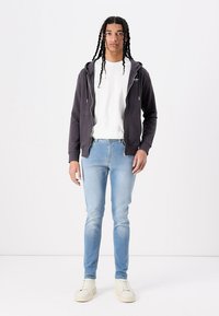 Sudadera con cremallera de color gris oscuro sobre una camiseta blanca, jeans de mezclilla azul claro y zapatillas beige. Las características incluyen un corte informal y puños acanalados.