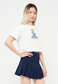 Un tricou scurt alb prezintă un grafic colorat cu flori și panglici. Este asociat cu o fustă mini plisată de un albastru închis. Textura materialului este netedă.