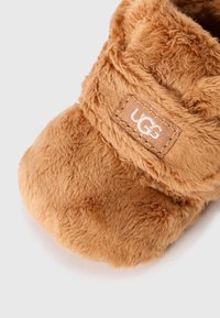 Pantoufle en fourrure tan avec une texture douce, bout arrondi et patch logo UGG. Le design présente un dessus replié pour plus de confort et de chaleur.