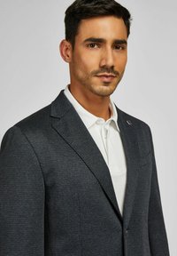 Mann mit rasiertem Bart, der einen dunkel strukturierten Blazer über einem weißen Hemd mit Kragen trägt, vor einem schlichten hellen Hintergrund.
