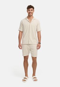 Helles beige Kurzarm-Hemd mit Knöpfen und passenden Shorts aus strukturiertem Stoff; getragen mit weißen Slipper-Sandalen.