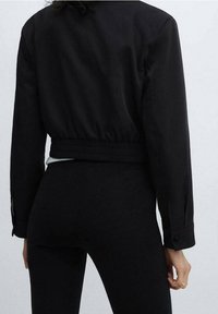 Veste noire courte avec ourlet élastique, manches longues et poignets à boutons. Tissu lisse avec finition mate et design minimaliste.