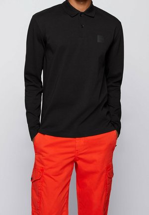 Mann trägt ein schwarzes langärmeliges Poloshirt mit zwei Knöpfen und leuchtend orangefarbene Cargohosen mit Seitentaschen, Hände in den Taschen.