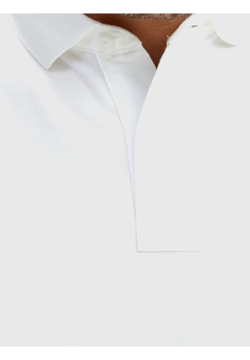Gros plan sur une personne portant un polo blanc à col avec le bouton du haut fermé.