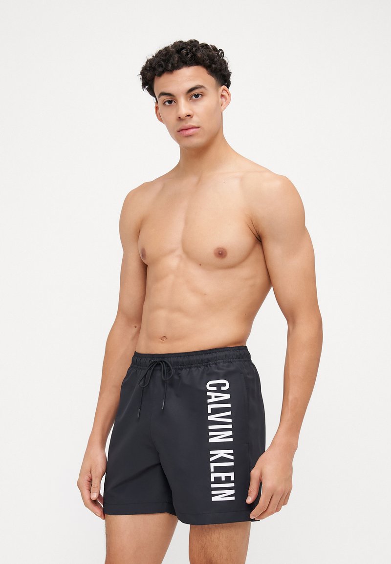 Shorts de bain noirs avec une taille à cordon, arborant un logo blanc "CALVIN KLEIN" verticalement sur le côté gauche. Tissu lisse et léger.