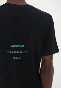 Melna kokvilnas T-krekls ar tirkīza izšuvumu, uz kura aizmugures ir teksts "JUSTCAVALLI", "Rock Your World" un "Milano".