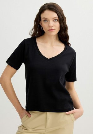 Mujer joven con cabello castaño rizado, lleva una camiseta negra de manga corta con cuello en V y pantalones beige, manos en los bolsillos, mirando hacia adelante.