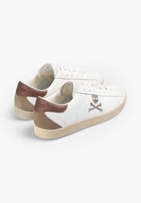 Sneakers bianchi in pelle con suola beige testurizzata, dettagli in suede marrone e glitter sul tallone, dotati di un logo con teschio e ossa incrociate.