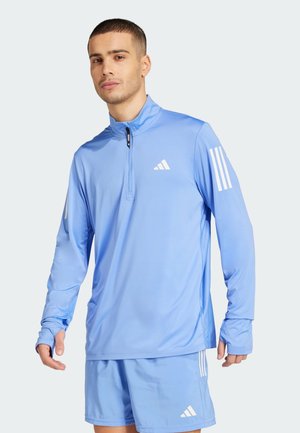 Maillot de sport à manches longues bleu clair avec un col zippé, en tissu évacuant l'humidité, accents blancs à trois rayures sur les manches et logo de la marque sur la poitrine.