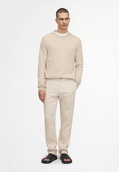 Man standing wearing beige long-sleeve sweater, beige pants, and black open-toe sandals against plain white background.