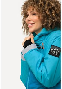 Giacca isolante teal con accenti grigi, polsini neri e un cordino arancione. Presenta un patch con logo di montagna sulla manica.