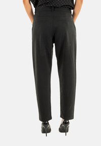 Pantalon noir au fini texturé, design à jambes fuselées et passants de ceinture. Porté avec des chaussures à talons hauts. Pas de poches visibles.
