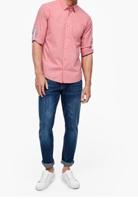 Camicia button-up rosa chiaro con piccoli pois blu, maniche arrotolate e taschino sul petto, abbinata a jeans slim-fit blu e sneakers bianche.
