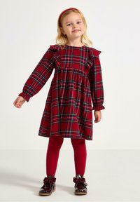 Rød ternet kjole med flæseaccenter, lange ærmer og elastisk talje; parret med røde leggings og blanke sorte sko med sløjfer.