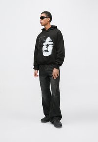 Črn prevelik pulover s kapuco z grafičnim potiskom obraza, kombiniran s temnimi jeans hlačami in temnimi čevlji, oblečen model z očali za sonce.