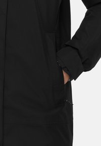 Giacca nera con finitura liscia e opaca, dotata di una tasca laterale e polsini regolabili. Il design presenta una silhouette pulita e minimalista.