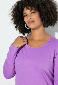 Dlouhý rukáv fialového topu s kulatým výstřihem, vyrobený z měkké, texturované látky; modelka pózuje s rukou na rameni, s minimem doplňků.