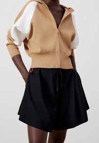 Brązowy kardigan zapinany na zamek z białymi rękawami, zestawiony z czarnymi szortami z regulowanym sznurkiem i czerwonym akcentem na pasku.