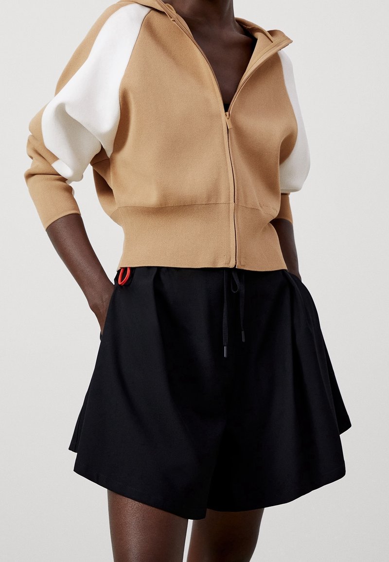 Brązowy kardigan zapinany na zamek z białymi rękawami, zestawiony z czarnymi szortami z regulowanym sznurkiem i czerwonym akcentem na pasku.