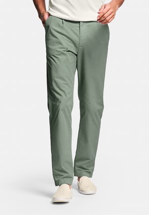 Homme portant un pantalon décontracté vert clair et des chaussures beiges à enfiler marchant devant un fond blanc uni.